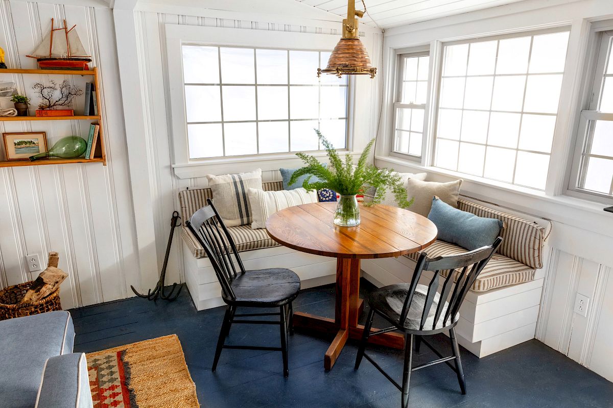 A cozy corner with a round wooden table, two chairs, a bench with cushions, and a plant in a vase. Natural light fills the room.