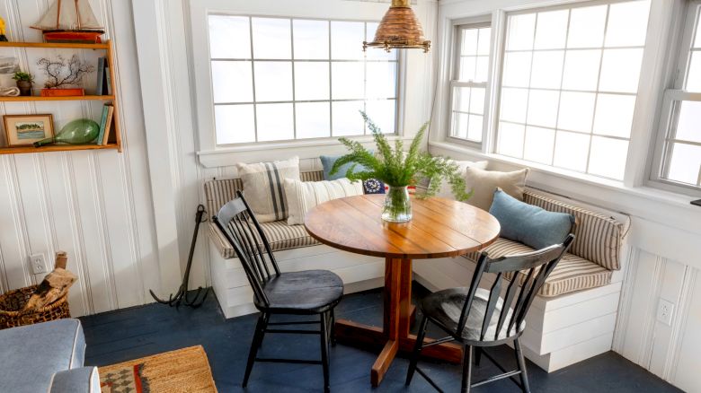 A cozy corner with a round wooden table, two chairs, a bench with cushions, and a plant in a vase. Natural light fills the room.