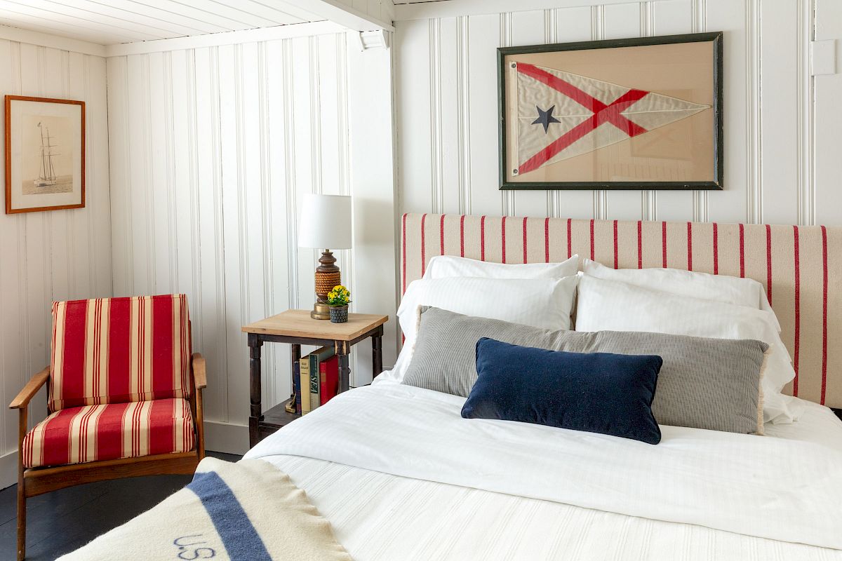 A cozy bedroom features a bed with striped red headboard and pillows, a striped chair, nautical decor, and framed art on white paneled walls.