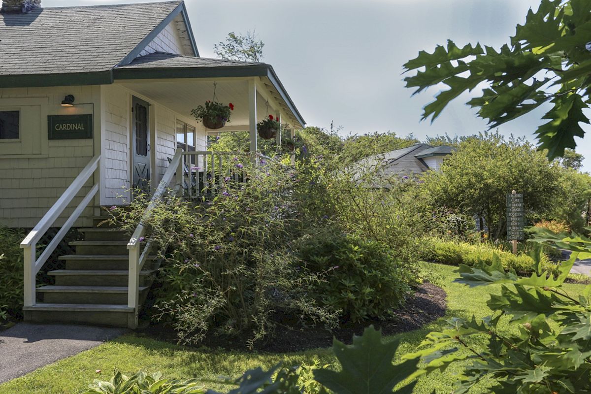 The image shows a quaint house with a porch, surrounded by lush greenery and plants. A small sign near the door reads