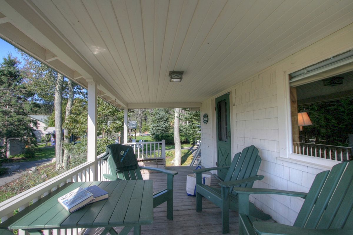 A cozy porch with green chairs and a table, an open book, and a view of the surrounding trees. A window and a door are on the right.
