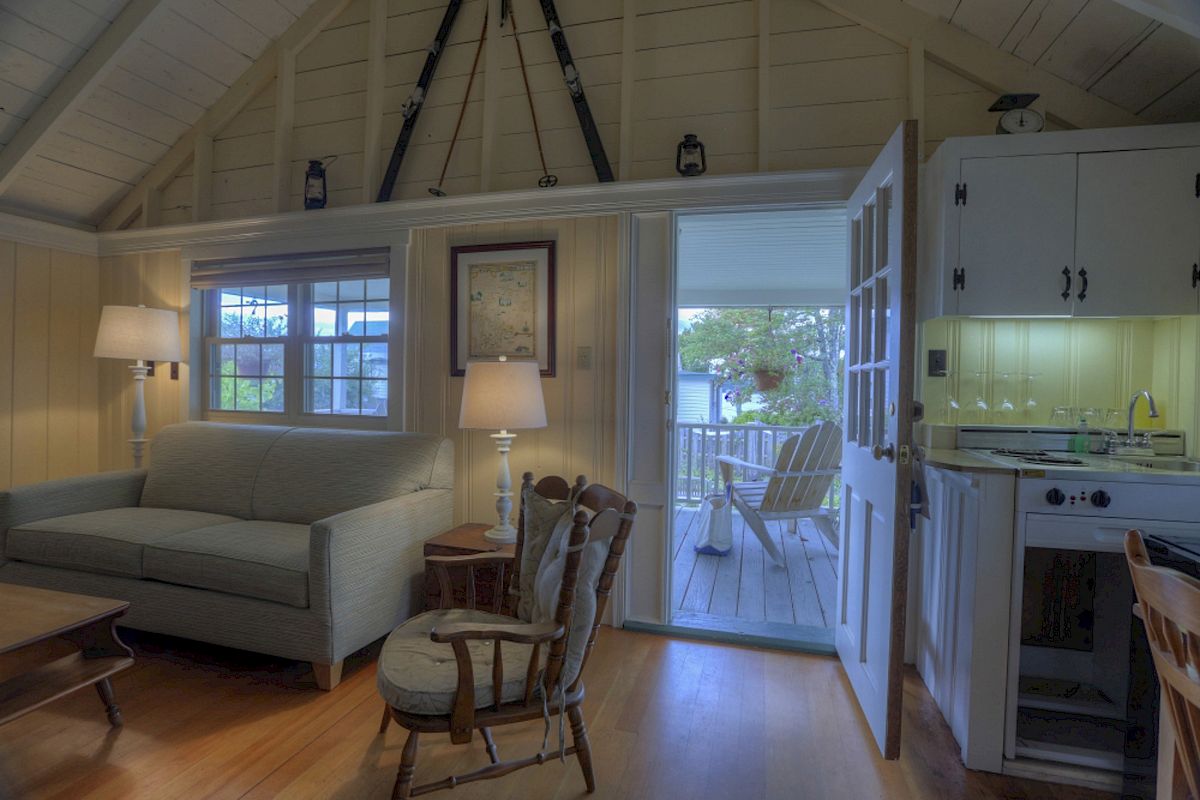 A cozy room has a sofa, rocking chair, lamps, and a kitchenette, opening to a veranda with Adirondack chairs.