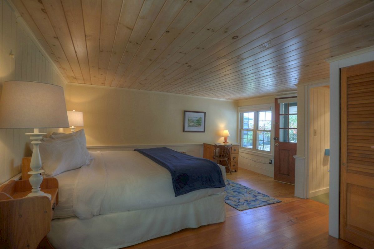 A cozy bedroom with wooden walls and ceiling, a large bed with white linens, a blue throw, a bedside lamp, a door, a rug, and a small desk.