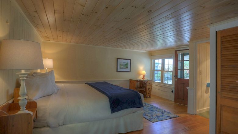 A cozy bedroom with wooden walls and ceiling, a large bed with white linens, a blue throw, a bedside lamp, a door, a rug, and a small desk.