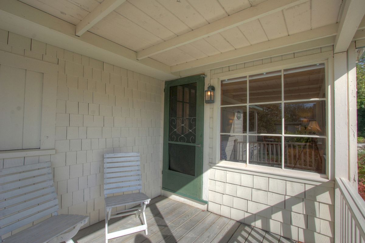 A porch with two white wooden chairs, a green door, and a large window with white framing. The area is shaded and has wooden flooring.