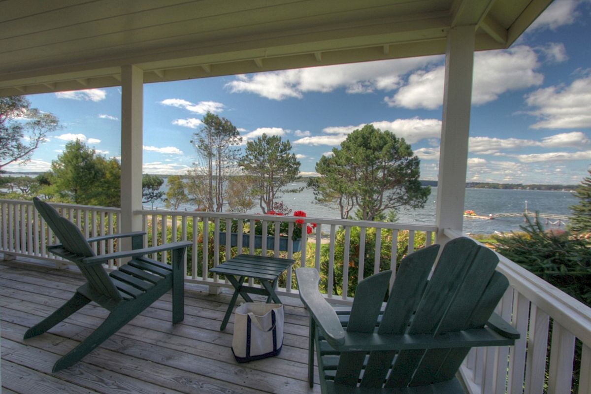 A porch with wooden chairs overlooks a scenic lake with trees. A small table holds a bag. The sky is partly cloudy, creating a serene atmosphere.