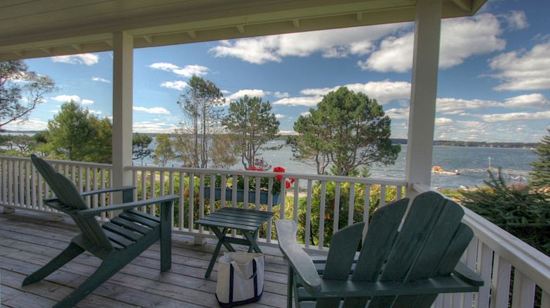 A porch with wooden chairs overlooks a scenic lake with trees. A small table holds a bag. The sky is partly cloudy, creating a serene atmosphere.