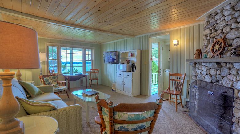 A cozy living room with wooden ceiling, stone fireplace, cushioned seating, a TV, and large windows letting in natural light, ending the sentence.