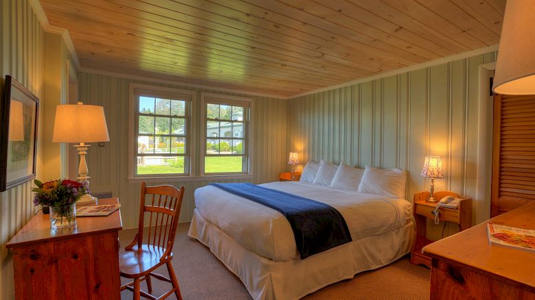 A cozy bedroom with a large bed, wooden furniture, and a desk. It has windows letting in natural light and is brightly lit with lamps.