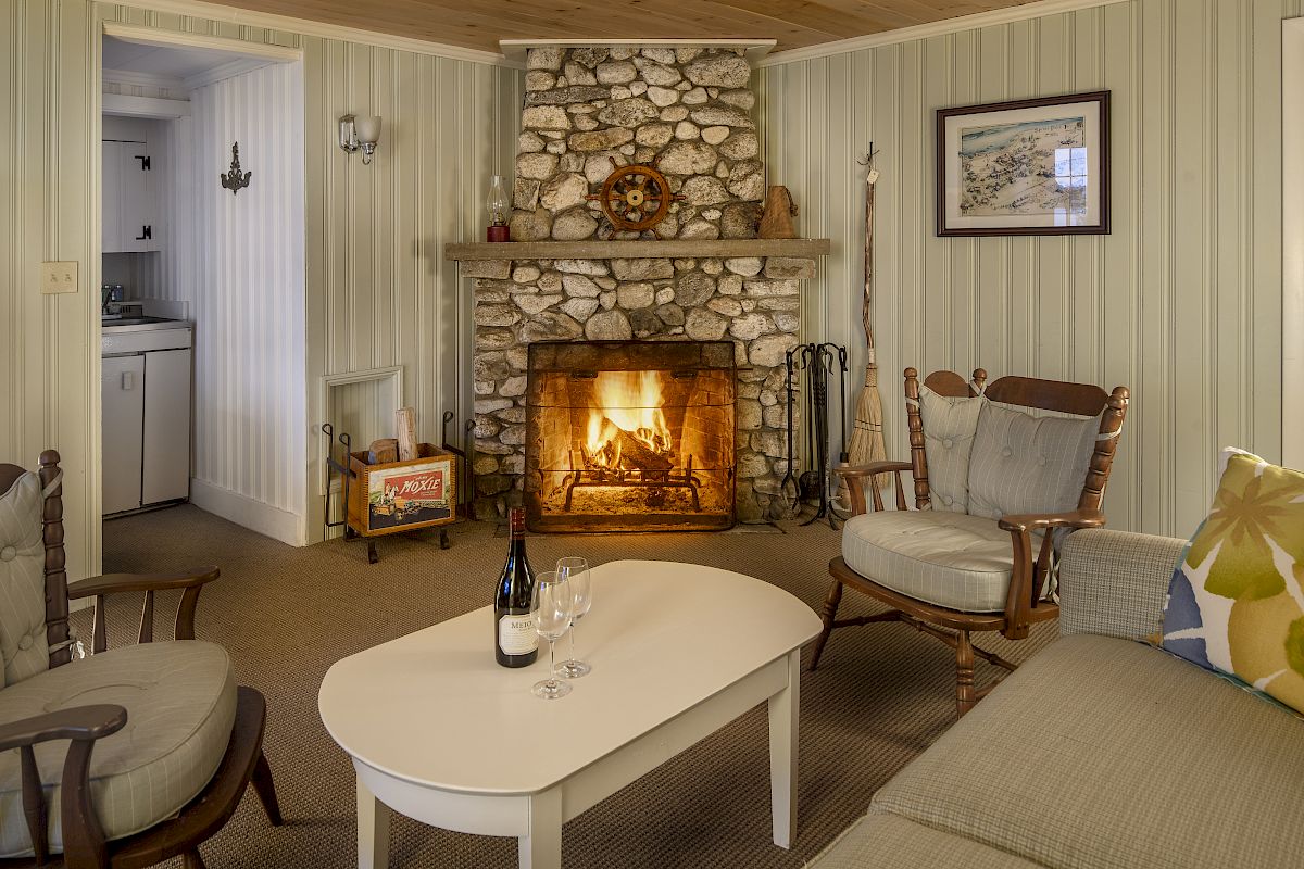 A cozy living room with a stone fireplace, two armchairs, a white coffee table with wine, and a sofa, leading to a small kitchen.