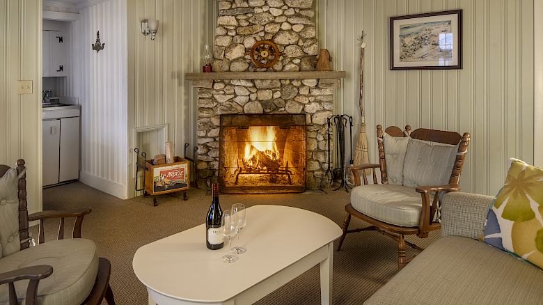 A cozy living room with a stone fireplace, two armchairs, a white coffee table with wine, and a sofa, leading to a small kitchen.