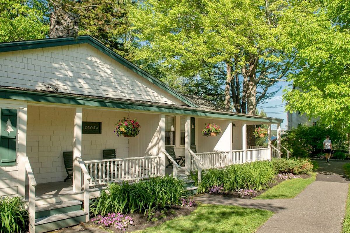 A cozy, white house with green trim is surrounded by lush greenery, hanging flower baskets, and a paved pathway under bright sunlight.