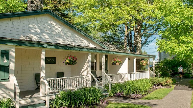 A cozy, white house with green trim is surrounded by lush greenery, hanging flower baskets, and a paved pathway under bright sunlight.