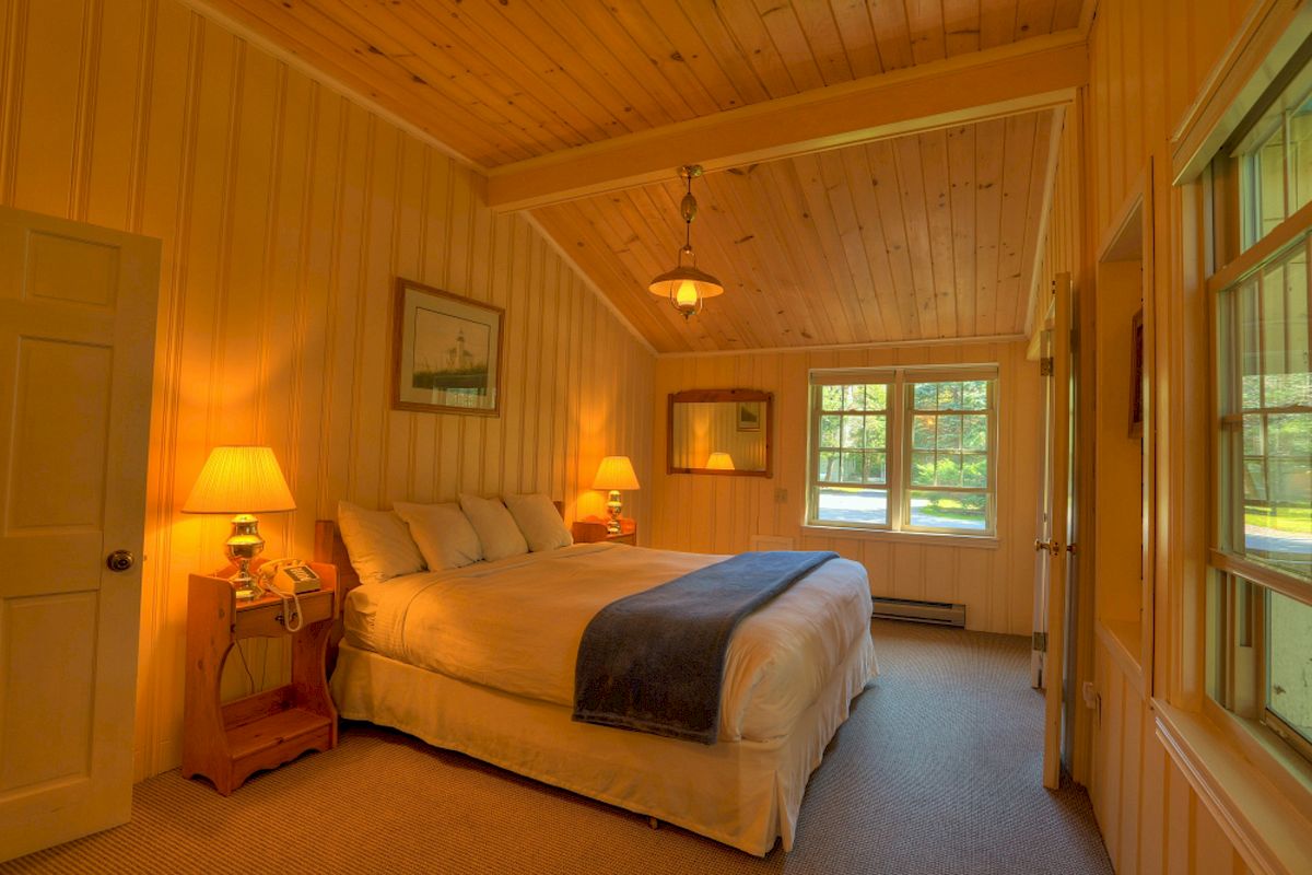 A cozy bedroom with wooden walls, a large bed, two nightstands with lamps, framed pictures, and windows letting in natural light.