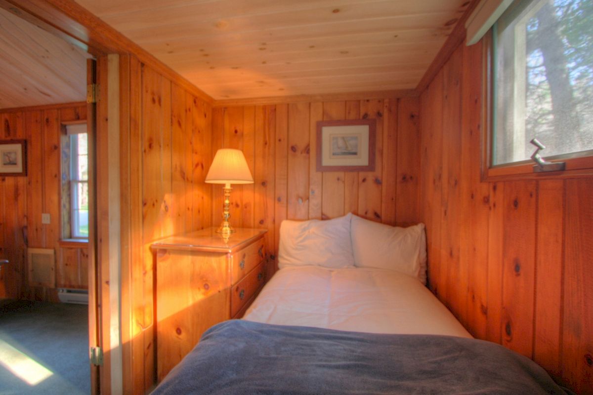 A cozy wooden room with a bed, lamp on a dresser, and picture frames on the wall. There is also a window letting in natural light.