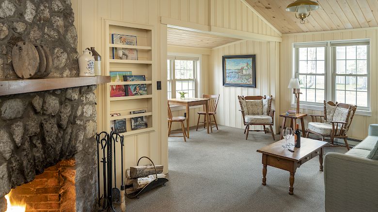 A cozy living room with a stone fireplace, bookshelves, seating area, a wooden coffee table, windows letting in natural light, and a small dining area.