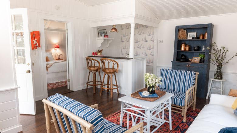 A cozy living room features wicker furniture, a small bar area, blue and white decor, artwork, and a view into a bedroom through an open doorway.