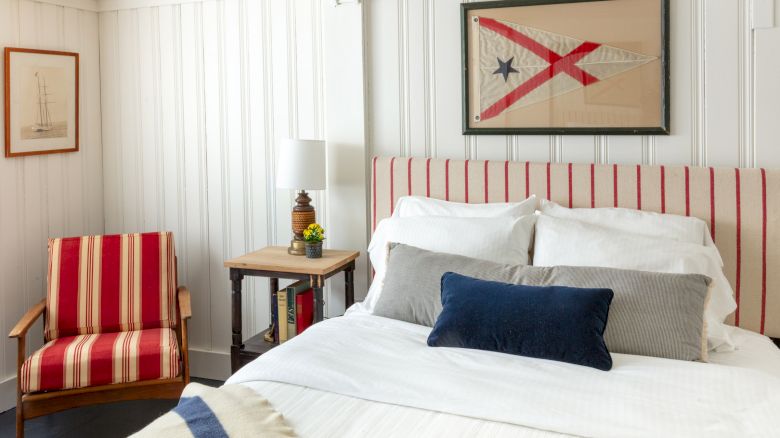 A cozy bedroom with a striped headboard and armchair, a bed with white and gray pillows, a flag artwork, and a small table with a lamp.