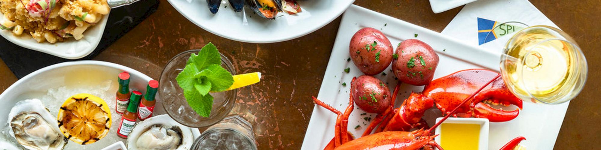 The image shows a variety of seafood dishes including lobster, oysters, mussels, and grilled bread, along with drinks and condiments on a table.