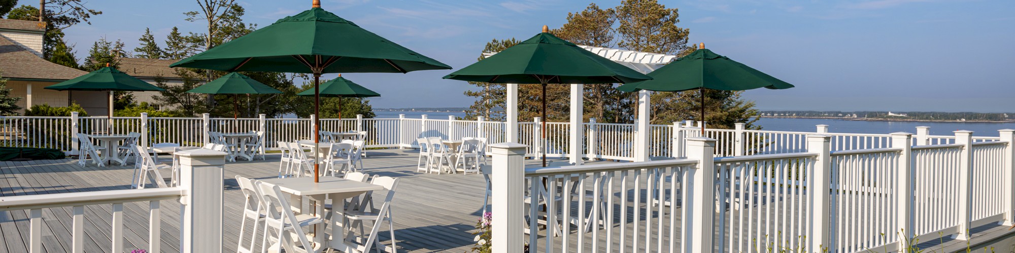 Outdoor deck with white railings, tables, and green umbrellas overlooking a scenic water view, surrounded by trees and flowers.