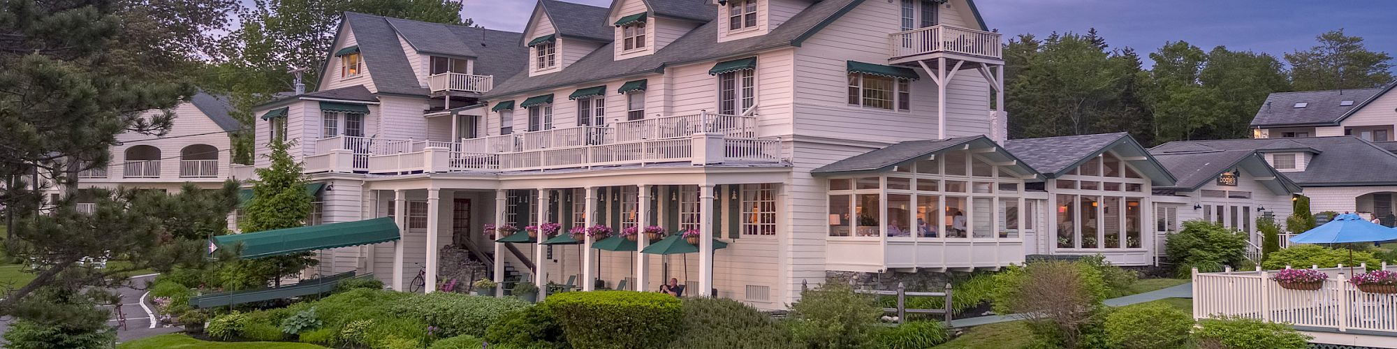 This image shows a large, multi-story house with a manicured lawn, white Adirondack chairs, and a backdrop of trees under a purple sky.