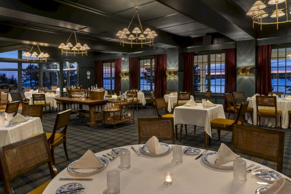 A dimly lit, elegant restaurant interior with neatly set tables, chandeliers, large windows, and a serene evening view outside the windows.