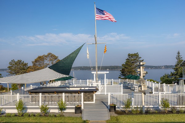 The image shows a deck with white railings, a large outdoor canopy, a flagpole with an American flag, and a water view surrounded by trees.