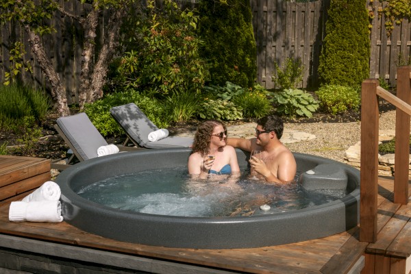 A couple relaxes in a hot tub surrounded by towels and lounge chairs, set in a backyard with greenery and a wooden fence.