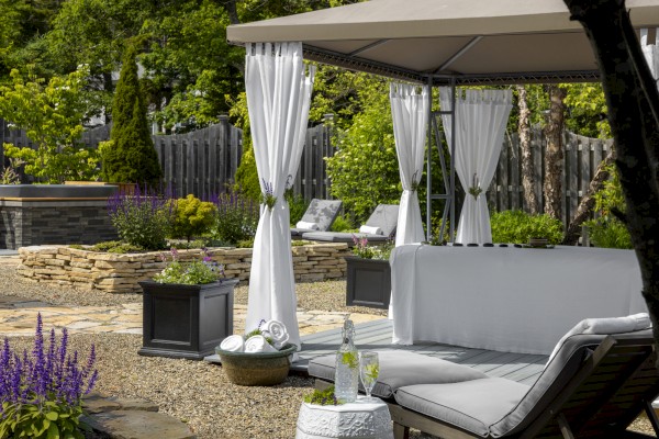 A tranquil outdoor seating area with a shaded gazebo, white curtains, loungers, potted plants, and a small table with water and towels.