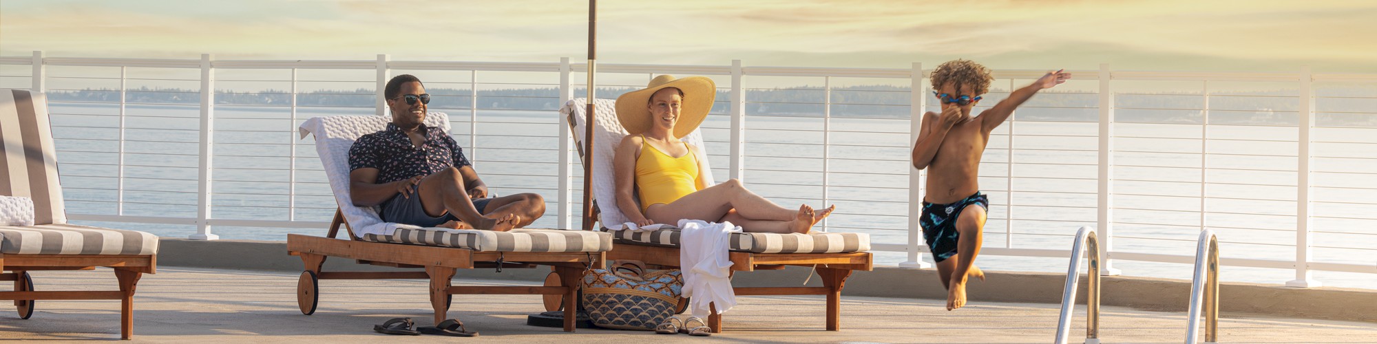 A family relaxing by a pool, with two adults on lounge chairs under umbrellas and two children playing in the water at sunset, is shown.