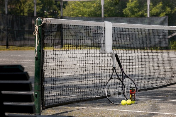 The image shows a tennis court with a net and two tennis rackets leaning against the net, along with a container of tennis balls and three loose tennis balls.