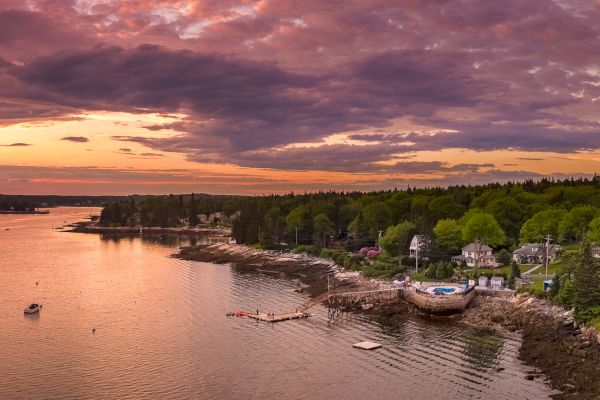 A serene coastal village at sunset with boats on the water, lush greenery, and a dramatic sky with hues of purple and orange ending the sentence.