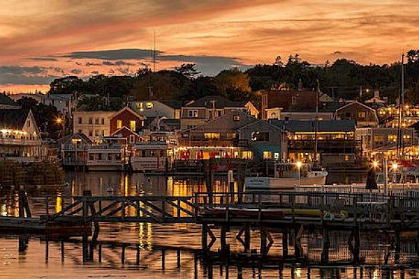 A picturesque harbor with a sunset sky, buildings illuminated along the waterfront, and boats docked on the calm water.