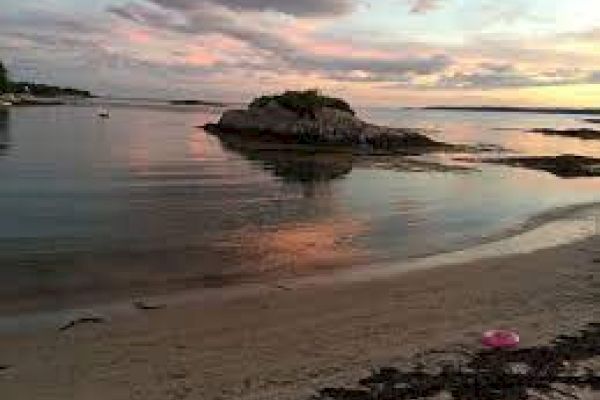 A sandy beach with some seaweed, calm water reflecting a colorful sunset, and a small island in the background, under a partly cloudy sky.