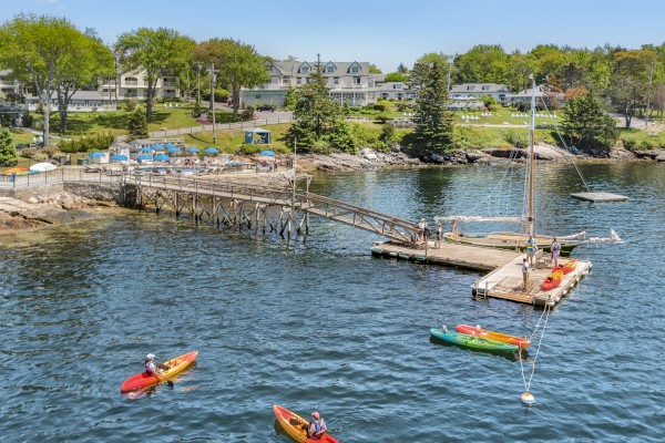 The image shows a scenic lakeside area with kayakers on the water, a dock with boats, and houses with lush greenery in the background, under a clear sky.