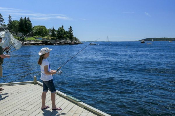 A man and a girl are fishing from a wooden dock on a sunny day with sailboats and an island visible in the background, ending the sentence.