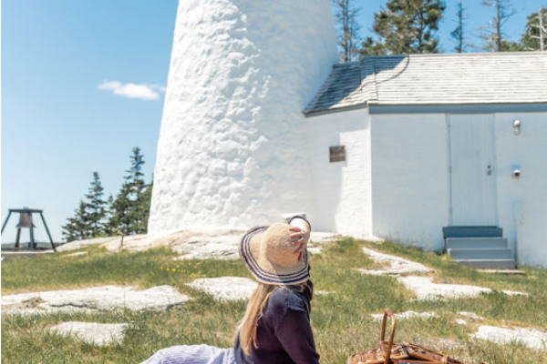 A woman sits on a picnic blanket with a basket in front of a white lighthouse on a sunny day.