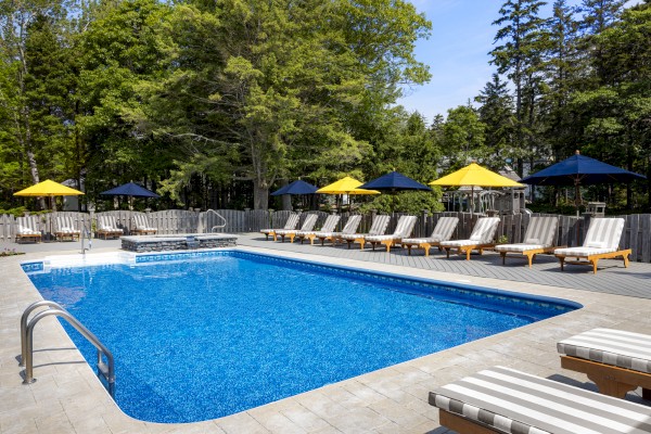 An outdoor pool surrounded by lounge chairs with striped cushions and umbrellas in a wooded area with trees in the background.