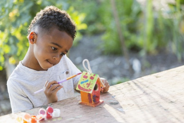 A young child is painting a small birdhouse outdoors with a brush and various colors of paint on a table.