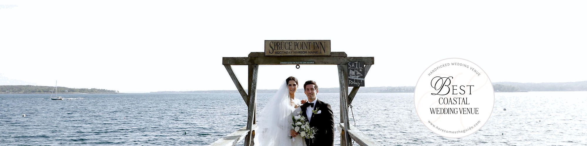 A bride and groom stand on a wooden dock by the water, with small boats visible in the background under a clear sky.