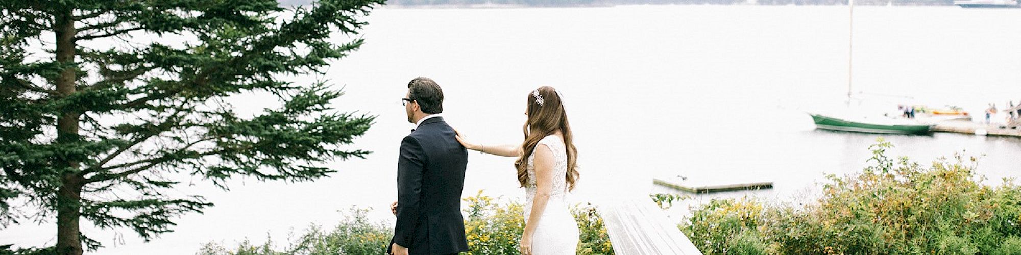 Two people by a lake, one in a wedding dress, trees nearby, serene