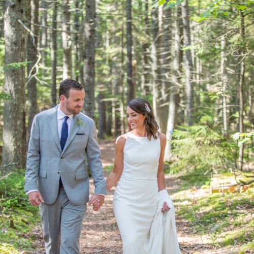 A couple in formal attire walks hand in hand through a lush forest, smiling and enjoying the natural surroundings.