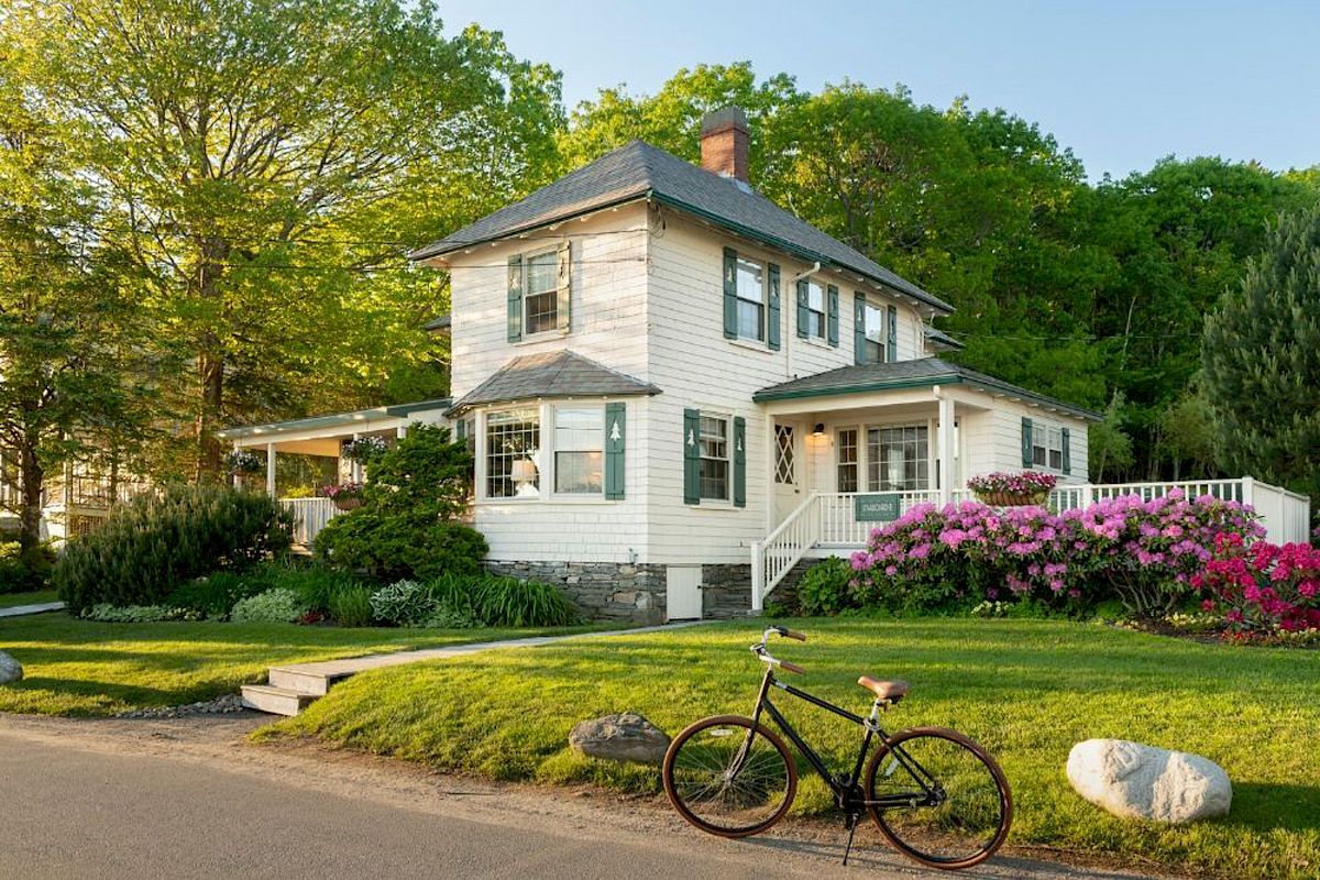 A charming two-story house with a bicycle in front, surrounded by vibrant greenery and colorful flowers, featuring a wrap-around porch.