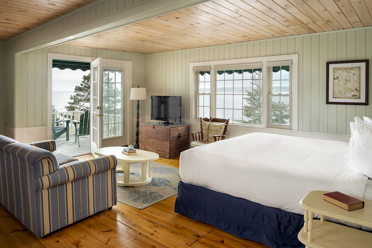 This image shows a cozy bedroom with a large bed, TV, sofa, and a view of the ocean from a balcony with outdoor seating.
