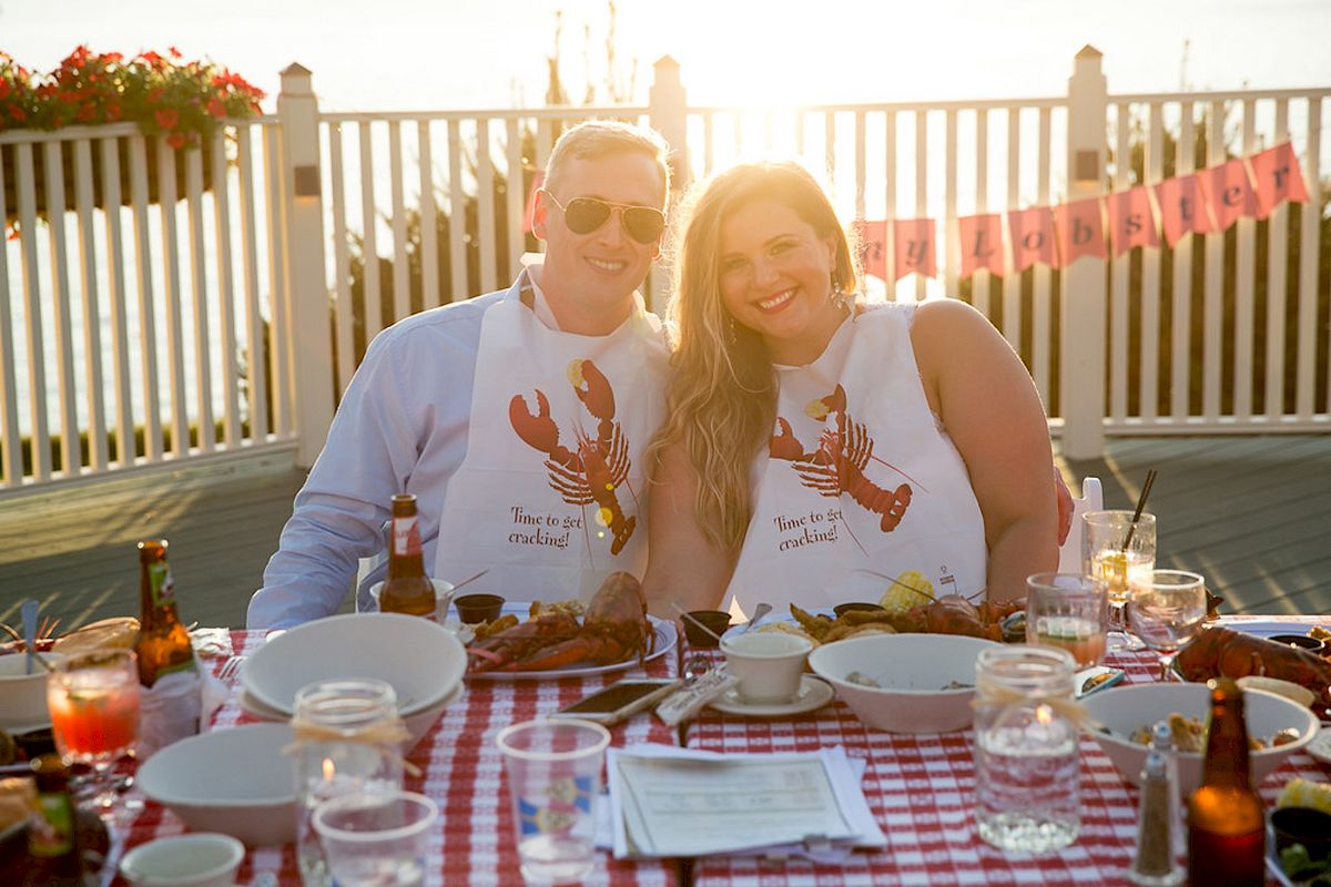 A couple wearing lobster bibs smiles at an outdoor table set for a meal, with drinks and food on a checkered tablecloth.