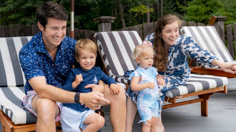 A family of four sits on lounge chairs outdoors; the parents and two young children are smiling, with the mother pointing at something.