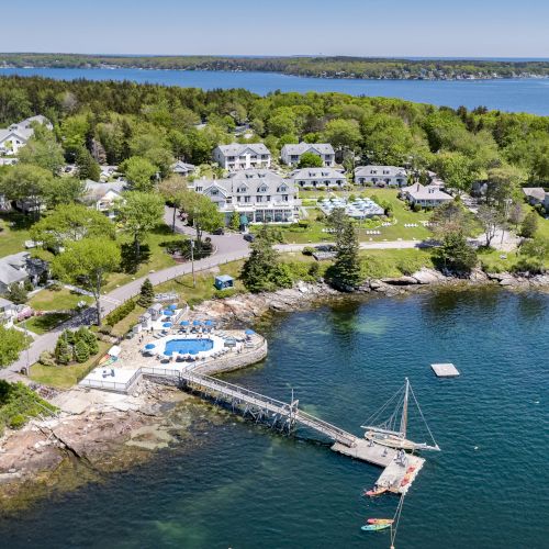 Aerial view of a waterfront resort with multiple buildings, a dock extending into the water, and a pool area surrounded by trees, ending the sentence.