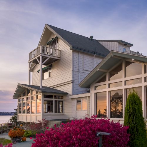 A large, modern house with expansive windows, surrounded by vibrant landscaping, overlooks a serene body of water at sunset.