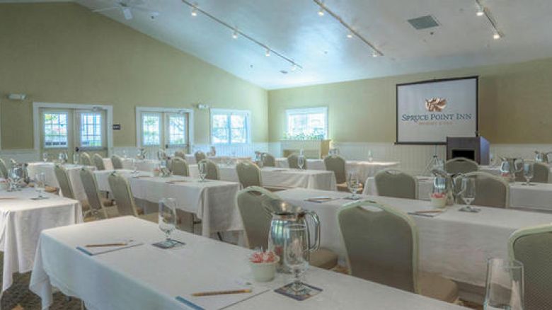 The image shows a conference room set up for a meeting or event with rows of tables and chairs, a screen displaying 