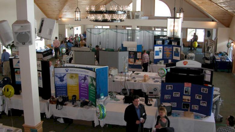 The image shows an indoor exhibition or trade show with various booths, informational displays, attendees, and some decorations like balloons.